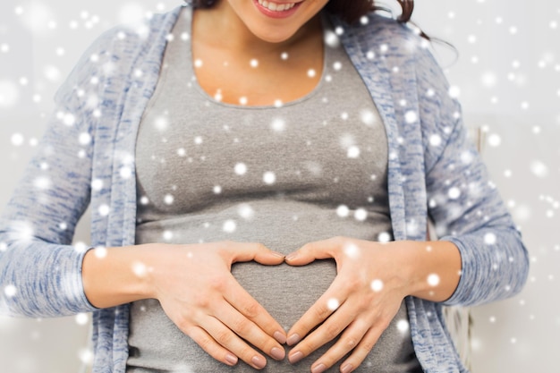 concept de grossesse, d'amour, de soins, de personnes et d'attentes - gros plan d'une femme enceinte heureuse faisant un geste cardiaque à la maison sur la neige