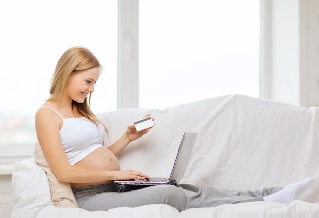 concept de grossesse, de maternité, d'argent, d'internet et de technologie - femme enceinte souriante assise sur un canapé avec un ordinateur portable et une carte de crédit