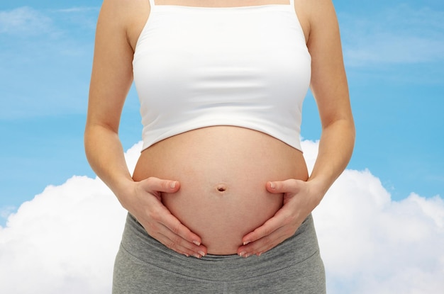 concept de grossesse, de maternité, de personnes et d'attente - gros plan d'une femme enceinte touchant son ventre nu sur fond bleu ciel et nuage