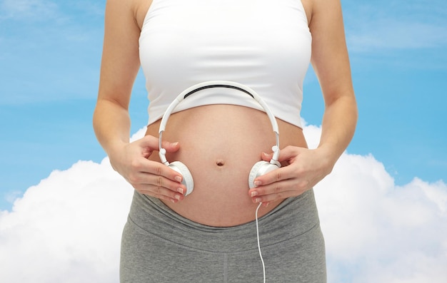 concept de grossesse, de personnes, de musique, de technologie et d'attente - gros plan d'une femme enceinte heureuse appliquant des écouteurs sur le ventre nu sur fond bleu ciel et nuage