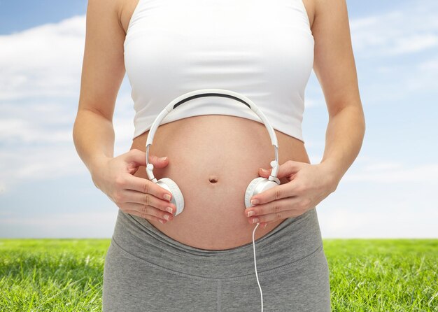 Photo concept de grossesse, de personnes, de musique, de technologie et d'attente - gros plan d'une femme enceinte heureuse appliquant des écouteurs sur le ventre nu sur fond de ciel bleu et d'herbe