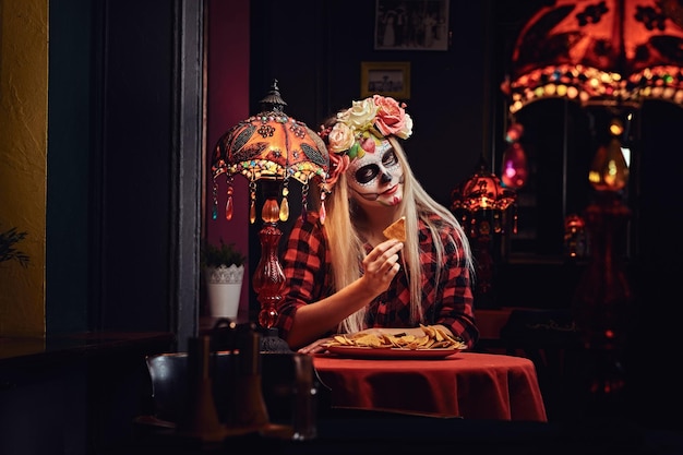 Concept d'Halloween et de Muertos. Jeune fille blonde avec un maquillage mort-vivant dans une couronne de fleurs mangeant des nachos dans un restaurant mexicain.