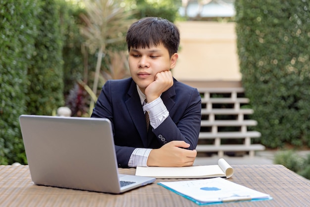 Concept d'homme d'affaires un jeune homme d'affaires en costume marine assis dans le jardin avec les documents et l'ordinateur portable sur la table, l'air réfléchi et sérieux.