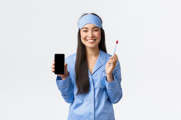 Concept d'hygiène, de technologie et de personnes à la maison. Jolie fille asiatique féminine en pyjama bleu et masque de sommeil tenant une brosse à dents, montrant un sourire blanc et un écran de smartphone, fond blanc