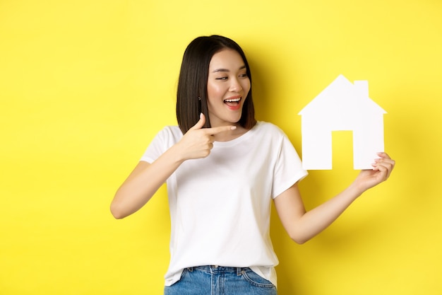 Concept immobilier et assurance. Joyeuse femme asiatique souriante, pointant et regardant la découpe de la maison en papier, recommande le logo de l'agence, debout sur fond jaune.