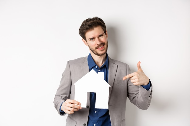 Concept Immobilier Et Assurance. Vendeur En Costume Gris Montrant La Découpe De Maison En Papier, Vente De Biens, Souriant à La Caméra, Fond Blanc.