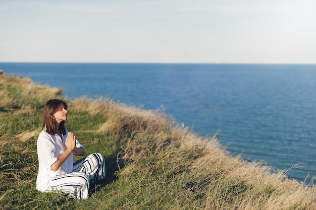 Concept de la journée internationale du yoga Jeune belle femme pratiquant le yoga sur la plage assise dans l'herbe Fille hipster faisant du yoga relaxant et écoutant les vagues de la mer Méditation calme bonheur