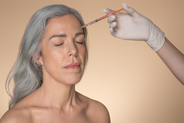 Concept de lifting de la peau, photo d'une femme âgée recevant une injection de produit de beauté dans le front beige