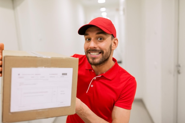 concept de livraison, de courrier, de personnes et d'expédition - homme heureux en uniforme rouge avec boîte à colis dans le couloir