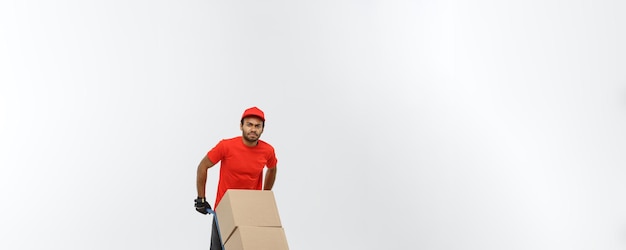 Concept de livraison Portrait d'un beau livreur afro-américain ou d'un coursier poussant un camion à main avec une pile de boîtes isolé sur fond de studio gris espace de copie