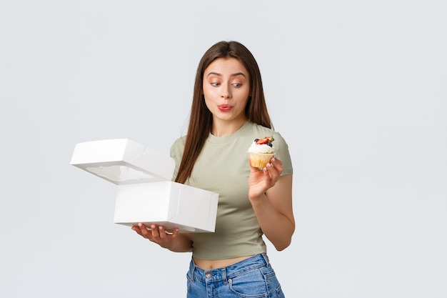 Concept de livraison, de style de vie et de nourriture. Une jolie femme idiote ouvre une boîte de desserts de pâtisserie ou de boulangerie, à la tentation et au désir de délicieux cupcake sucré, fond blanc