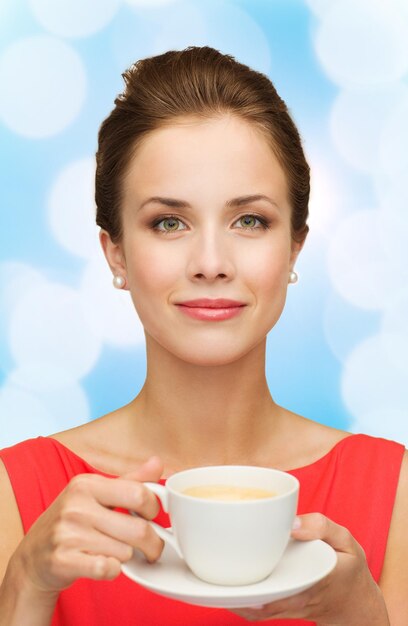 Concept de loisirs, de bonheur et de boisson - femme souriante en robe rouge avec une tasse de café sur fond de lumières bleues