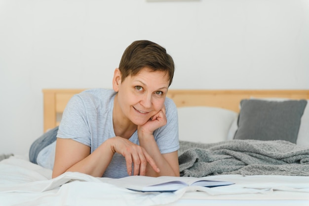 Concept de loisirs et de maison souriante femme d'âge moyen lisant un livre et allongée sur un canapé à la maison