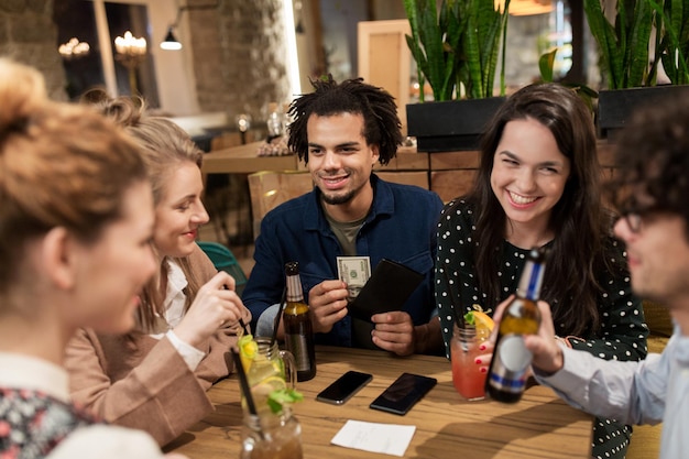 concept loisirs, personnes et vacances - amis heureux avec boissons, argent et facture au bar ou au café