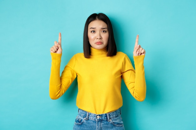 Concept de magasinage. Femme asiatique déçue et sombre bouder bouleversé, pointant du doigt la bannière de mauvaises nouvelles, debout sur le bleu.