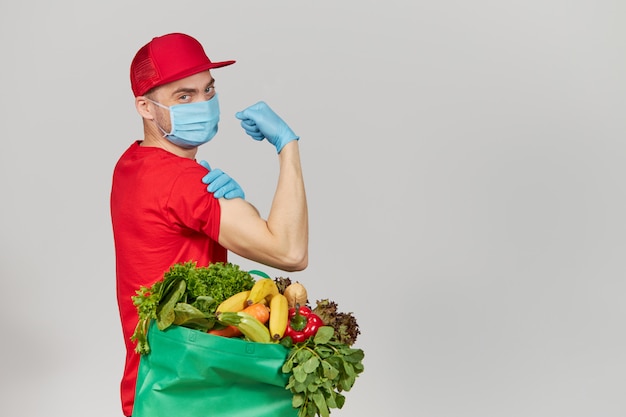 Concept De Magasinage En Ligne. Courrier Masculin En Uniforme Rouge, Masque De Protection Et Gants Avec Une Boîte D'épicerie Avec Des Fruits Et Légumes Frais. Livraison à Domicile De Nourriture Pendant La Mise En Quarantaine Du Coronavirus