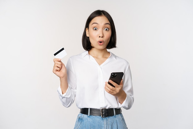 Concept de magasinage en ligne Image d'une fille asiatique surprise tenant une carte de crédit et un smartphone à la recherche d'incrédulité face à l'arrière-plan blanc de l'appareil photo