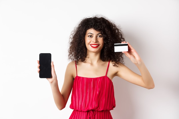 Concept de magasinage en ligne. Modèle féminin élégant aux cheveux bouclés, vêtu d'une robe rouge, montrant une carte de crédit en plastique avec écran de téléphone portable vide, debout sur fond blanc.