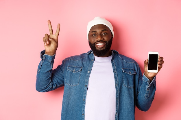 Concept de magasinage en ligne et de technologie. Heureux gars noir en bonnet et chemise en jean montrant un écran mobile et un signe de paix, debout sur fond rose