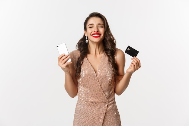 Concept de magasinage en ligne et de vacances. Femme satisfaite et heureuse en robe élégante souriante, utilisant une carte de crédit et un téléphone portable, debout sur fond blanc.
