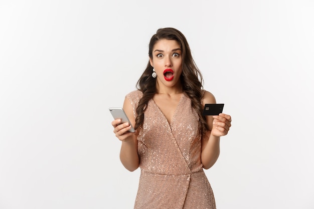Concept de magasinage en ligne et de vacances. Femme surprise en robe glamour, achetant sur Internet avec carte de crédit et téléphone portable, l'air étonné, fond blanc.