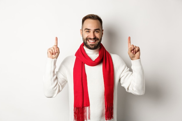 Concept de magasinage de Noël et de vacances d'hiver. Homme souriant avec barbe montrant le logo, pointant les doigts vers le haut et l'air heureux, debout dans une écharpe rouge avec un pull, fond blanc.