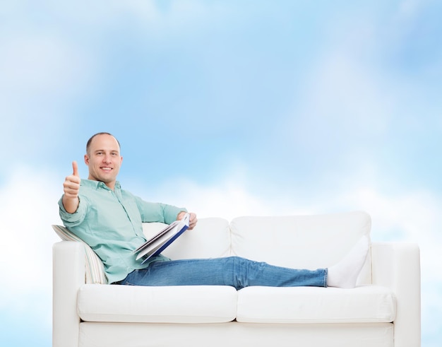 concept de maison, de loisirs et de bonheur - homme souriant allongé sur un canapé avec un livre et montrant les pouces vers le haut