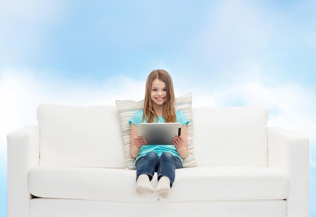 concept de maison, de loisirs, de technologie et de bonheur - petite fille souriante assise sur un canapé avec un ordinateur tablette