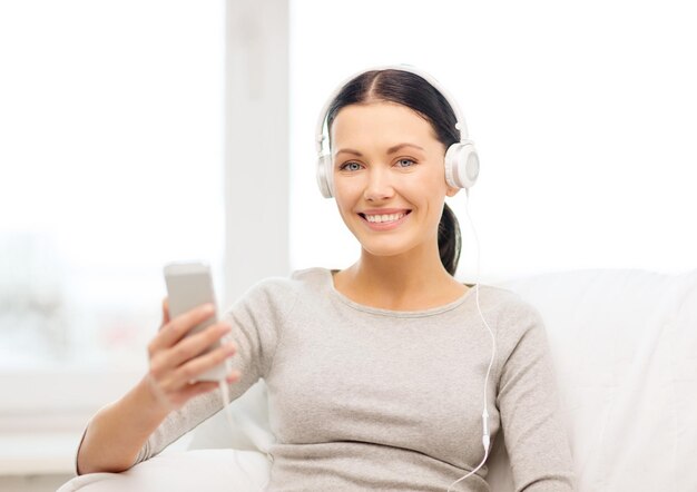 concept de maison, de technologie, de communication et d'internet - femme assise sur le canapé avec smartphone et casque à la maison