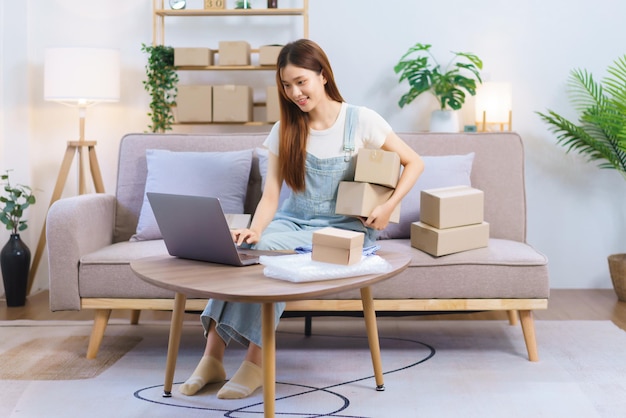Concept de marchand en ligne Une femme entrepreneur détient des boîtes à colis et vérifie les commandes en ligne sur un ordinateur portable