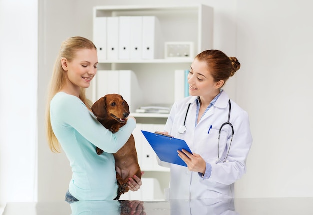 concept de médecine, de soins aux animaux de compagnie et de personnes - femme heureuse tenant un chien teckel et un médecin vétérinaire avec presse-papiers à la clinique vétérinaire