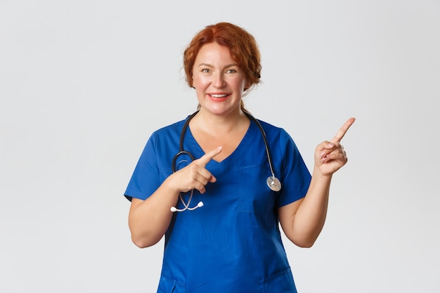 Concept de médecine, de soins de santé et de coronavirus. Impressionné et excité, mignon médecin rousse d'âge moyen, infirmière en gommage montrant la voie, pointant du doigt le coin supérieur droit amusé avec une offre impressionnante.