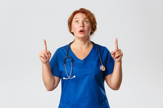 Concept de médecine, de soins de santé et de coronavirus. Médecin d'âge moyen rousse impressionnée et excitée, infirmière en gommage perdant la parole d'étonnement en voyant quelque chose de génial, pointant du doigt vers le haut.