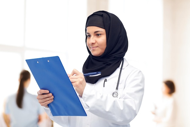 concept de médecine, de soins de santé et de personnes - une médecin musulmane souriante portant un hijab et un manteau blanc avec un bloc-notes et un stéthoscope sur le fond de la clinique
