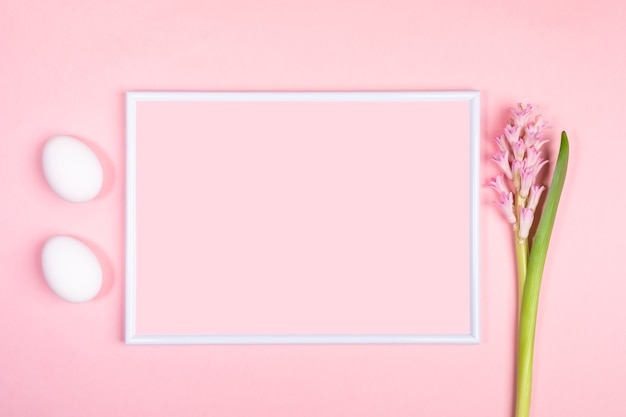 Concept minimal de fête de Pâques avec cadre blanc, deux œufs blancs et fleur de jacinthe sur fond rose pastel.