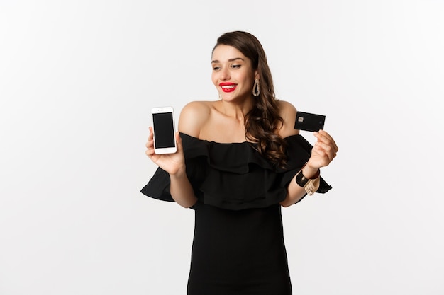 Concept de mode et de magasinage en ligne. Heureuse jeune femme en robe noire, montrant une carte de crédit et un écran mobile, debout sur fond blanc