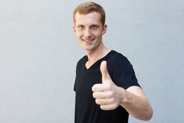 Concept de mode, de style, d'émotions et de personnes - Portrait en gros plan d'une rousse d'un beau mec viril avec des taches de rousseur sur un T-shirt noir à fond gris. montrant des coups et souriant joyeusement