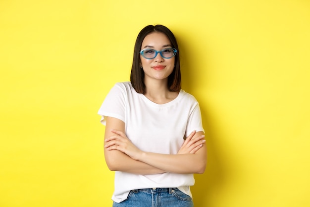 Concept de mode et de style de vie. Femme asiatique impertinente et confiante dans des lunettes de soleil à la mode à la recherche de confiance en soi à la caméra, debout sur jaune.