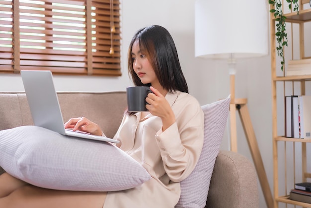 Concept de mode de vie à la maison Jeune femme asiatique surfant sur les médias sociaux sur un ordinateur portable et buvant du café