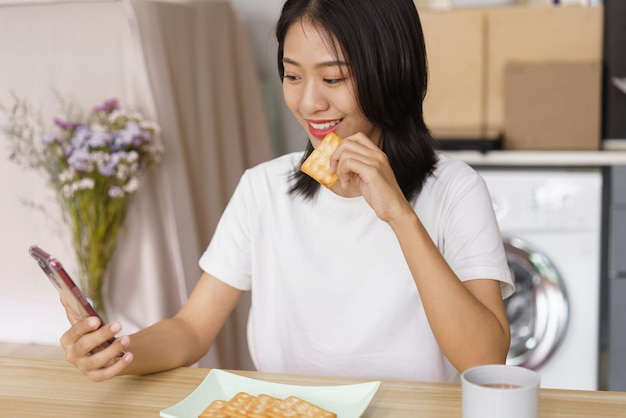 Concept de mode de vie à la maison Jeune femme mangeant des craquelins et surfant sur les réseaux sociaux sur smartphone à la maison