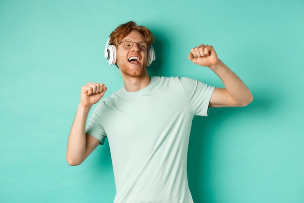 Concept de mode de vie. Un mec insouciant aux cheveux roux, dansant et s'amusant, écoutant de la musique dans des écouteurs, debout sur fond turquoise.