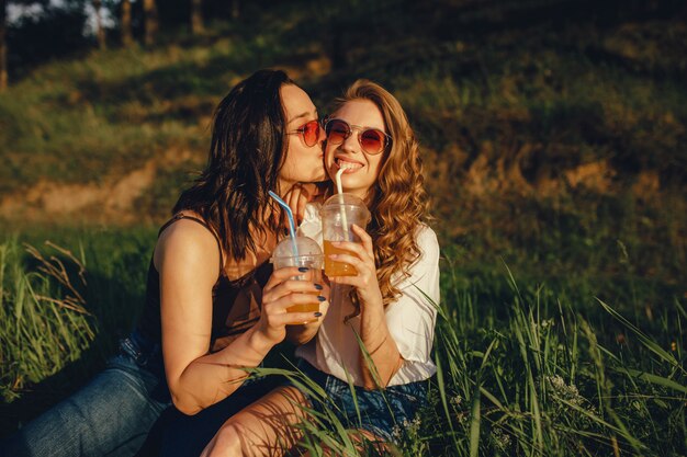 Concept de mode de vie: portrait de filles heureuses s'amuser, s'asseoir sur l'herbe, s'embrasser sur la joue, boire un cocktail à lunettes de soleil, au coucher du soleil, expression faciale positive, en plein air