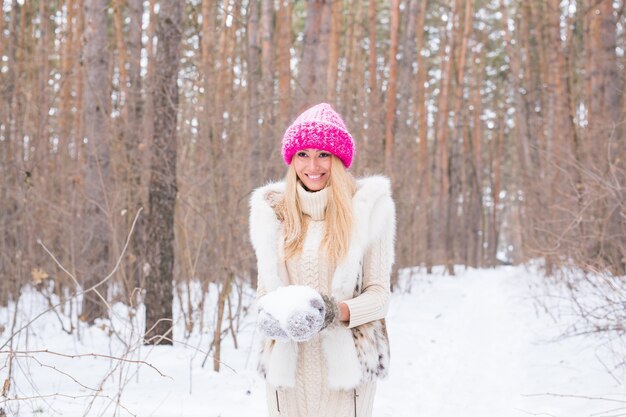 Concept de nature et de personnes - jolie femme blonde habillée en manteau et chapeau rose dans le parc d'hiver tenant la neige dans ses mains.