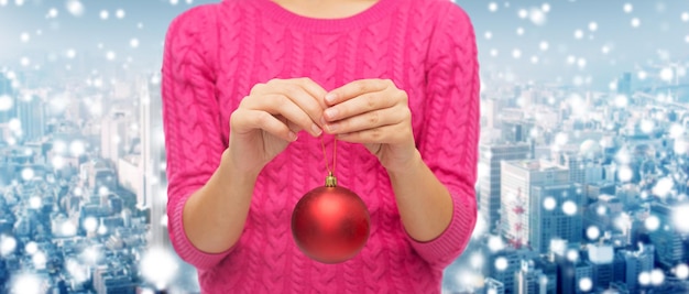 concept de noël, de décoration, de vacances et de personnes - gros plan d'une femme en pull rose tenant une boule de noël sur fond de ville enneigée