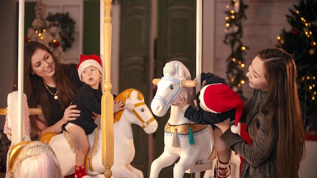 Concept de Noël deux bébés assis sur des chevaux de carrousel à l'intérieur et jouant avec eux leur mère