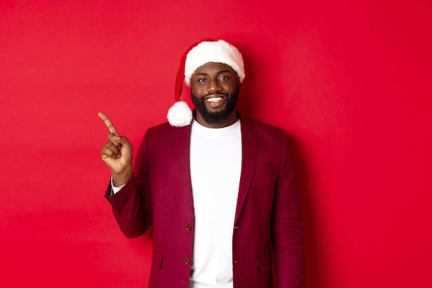 Concept de Noël, fête et vacances. Bel homme noir en bonnet de noel pointant le doigt vers la gauche, montrant une publicité, debout heureux sur fond rouge
