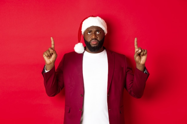 Concept de Noël, fête et vacances. Un homme afro-américain bouleversé en bonnet de noel se plaignant, pointant les doigts vers le haut et fronçant les sourcils déçu, debout sur fond rouge