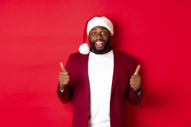 Concept de Noël, fête et vacances. Joyeux homme noir en bonnet de noel montrant le pouce levé, célébrant le Nouvel An, approuvant et acceptant, debout sur fond rouge
