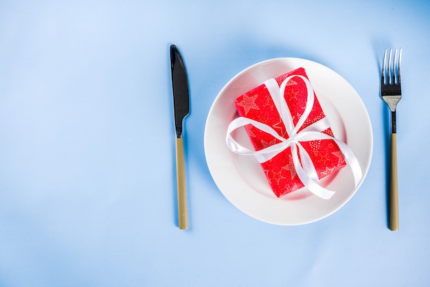 Concept de Noël, fond de salle à manger avec assiette, fourchette, couteau et boîte-cadeau dans un emballage festif, espace de copie de fond bleu clair au-dessus