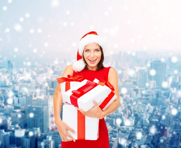 concept de noël, d'hiver, de bonheur, de vacances et de personnes - femme souriante en bonnet d'assistance avec des coffrets cadeaux sur fond de ville enneigée
